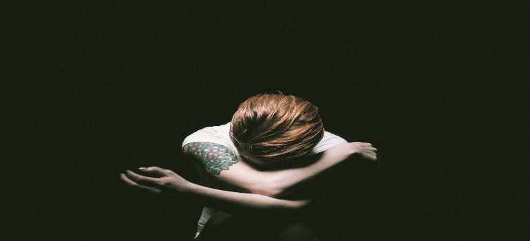 A person with a tattoo hugs their knees, bowing their head in a dark space, conveying a sense of vulnerability and introspection.